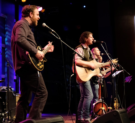 three men singing playing guitar on stage